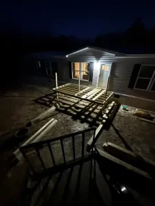 A wooden deck under the lights at night. The home connected to it is quaint and can faintly be seen through the orange window lighting.